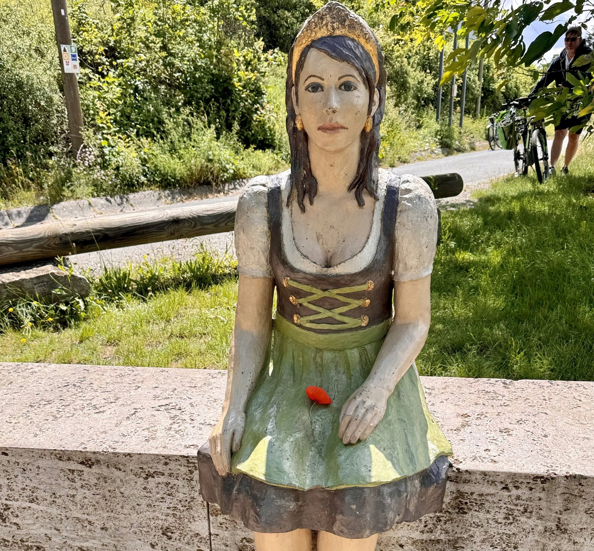Die lebensgroße Figur aus Tonerdeschmelzzement stellt eine Weinprinzessin dar, die auf einer steinernen Bank sitzt. 